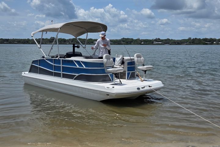 Pontoon boat anchored on sandy shore with person onboard, cloudy sky.