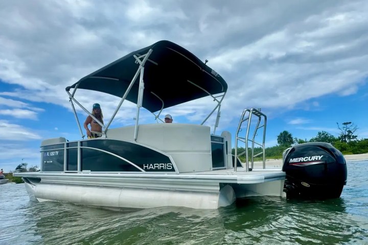 Pontoon boat with canopy on water, two people onboard, trees and cloudy sky in background.