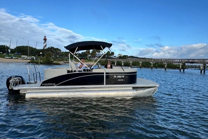 Pontoon boat on water with two people, lighthouse in background under blue sky.