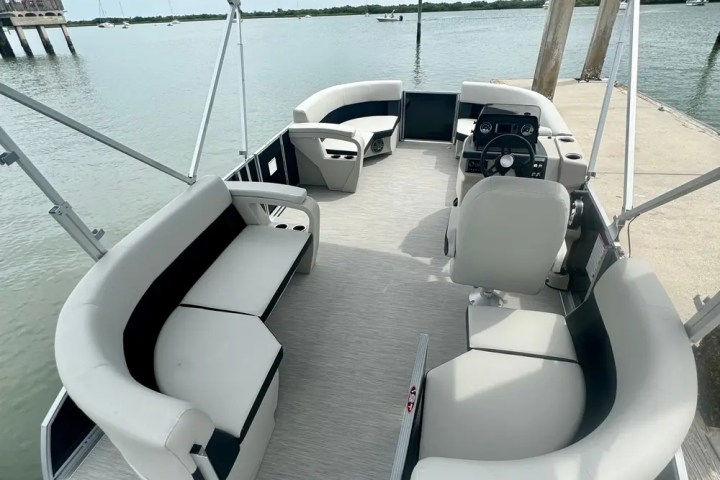 Pontoon boat interior with seating, steering wheel, and canopy, docked on a calm body of water.