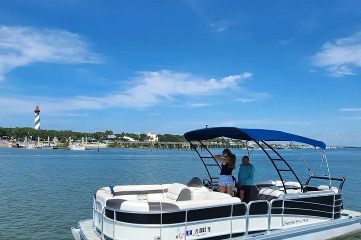 Pontoon boat on water with people, lighthouse and marina in background, blue sky overhead.