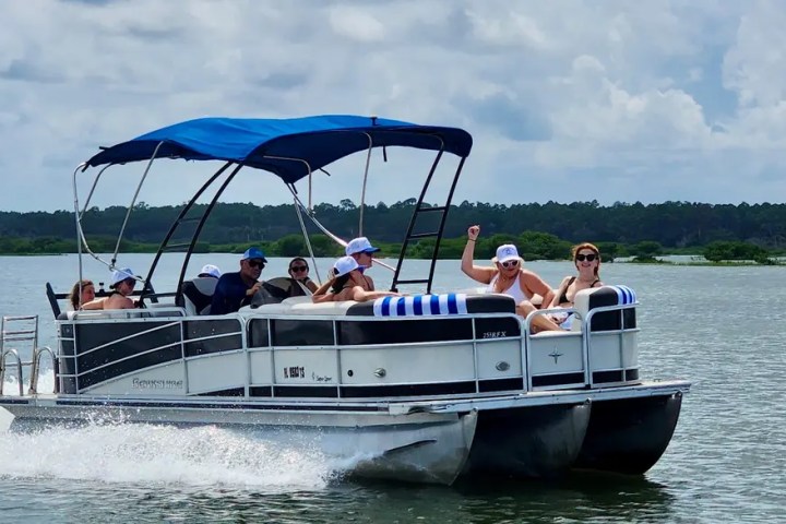 People on a pontoon boat with a blue canopy cruising on a lake under a cloudy sky.