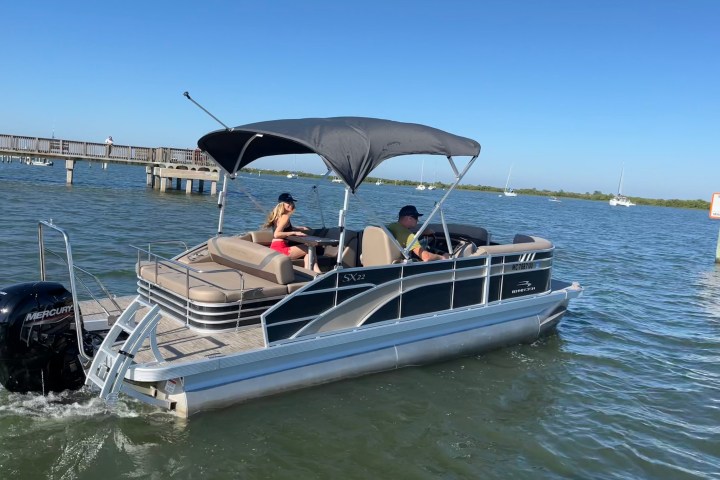 Pontoon boat with canopy on water, two people seated, clear sky, distant boats and pier visible.