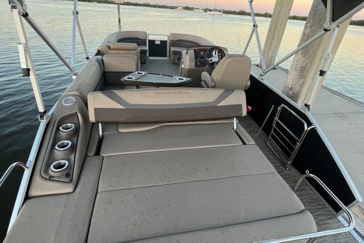 Interior view of a pontoon boat with seats, table, and canopy, docked at a lake during sunset.