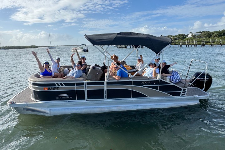 Group of people on a pontoon boat waving, under a clear blue sky.
