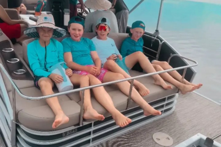 Four children in hats sitting on a boat sofa near water, wearing teal shirts and shorts, smiling.
