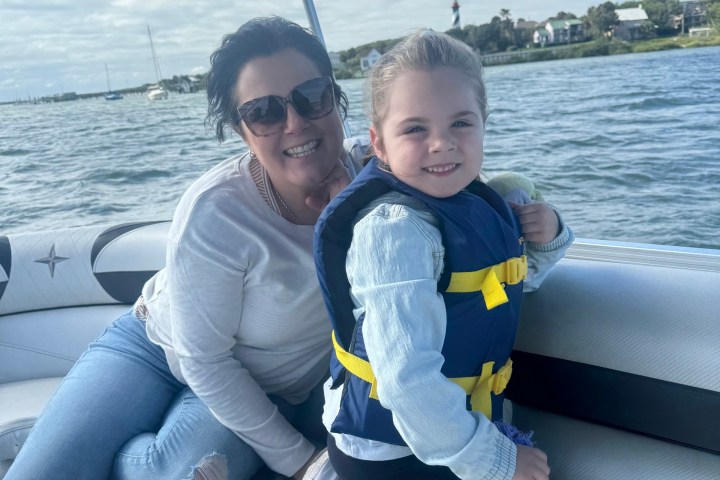 Woman and child in life vest on a boat, smiling with water and cloudy sky in background.