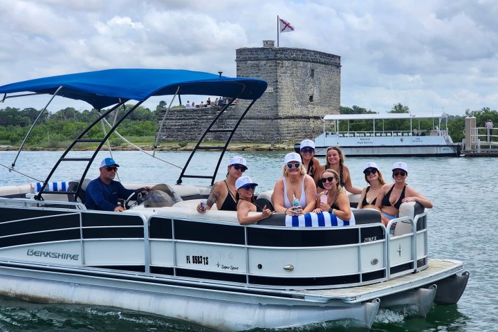 Group on a pontoon boat with stone building and flag in background on a cloudy day.