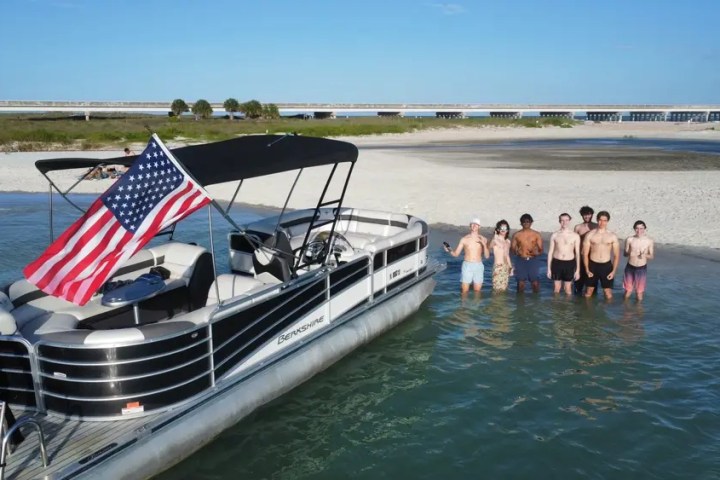 A pontoon boat with an American flag and a group of people standing in shallow water on a sunny day.