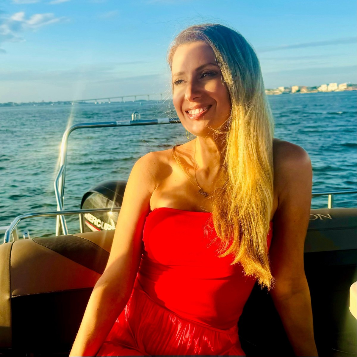 Woman in a red dress smiling on a boat with ocean and bridge in background.