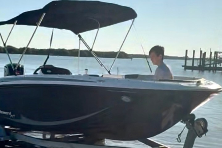 Person steering a boat with a canopy on a trailer by the water.