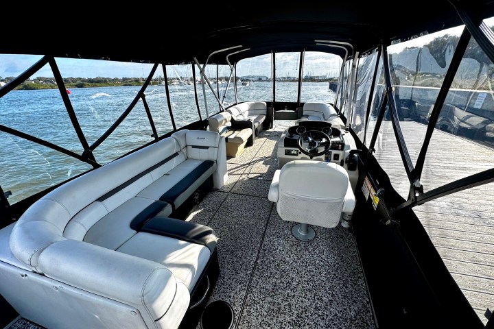 Interior of a boat with white seating and a steering wheel, surrounded by water and docks.