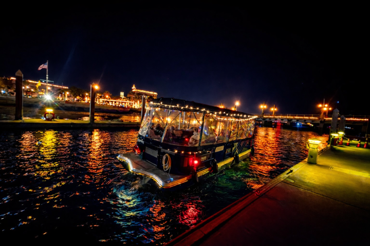 A boat with string lights on a river at night, with a lit-up waterfront and bridge in the background.