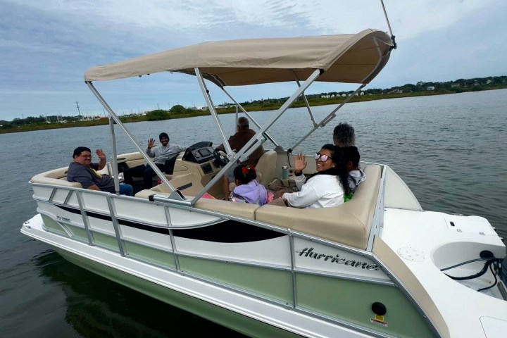 Six people on a pontoon boat with a canopy, waving on a calm lake.