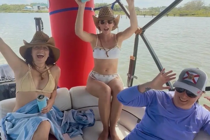 Three people on a boat, two women in hats, one woman in a bikini, enjoying a sunny day on the water.