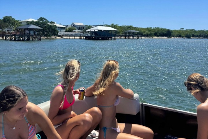 Four people on a boat in swimsuits, looking at waterfront houses on a sunny day.
