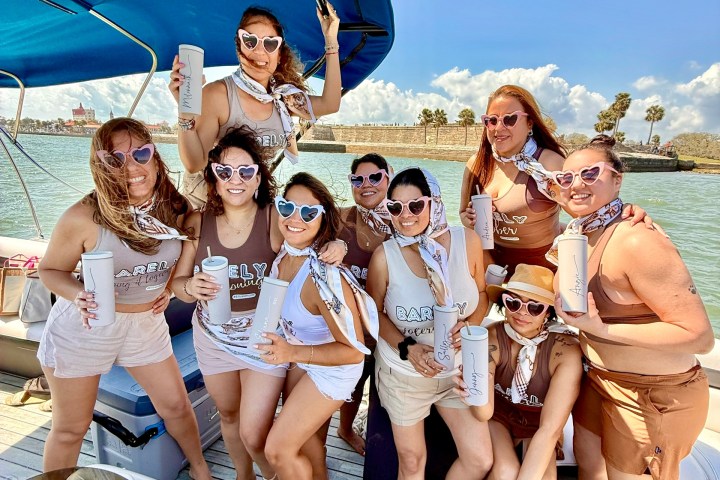 Group of women in heart sunglasses on a boat holding cups, smiling under a sunny sky.
