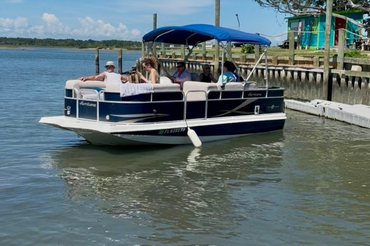 A pontoon boat with people on a calm river near a dock.